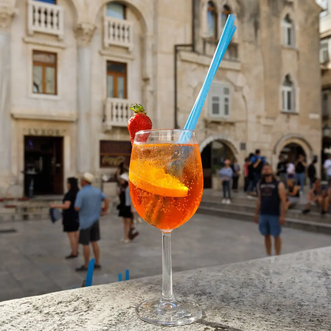 An Aperol Spritz with a strawberry on the stone ledge outside Lvxor, Peristil Square behind